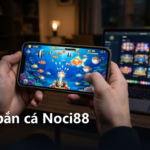 Người chơi cầm điện thoại bắn cá, phía sau là laptop mở sảnh game Noci88
